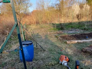 Vue réelle en Moselle d'une débroussailleuse au premier plan, devant un potager en préparation. Sur le côté droit, des planches de culture déjà surélevées avec des rondins de bois et du terreau noir sont prêtes pour la plantation. Cette photo illustre concrètement comment installer un petit potager débutant sans bêcher, en gérant la végétation existante avant de créer une zone fertile.