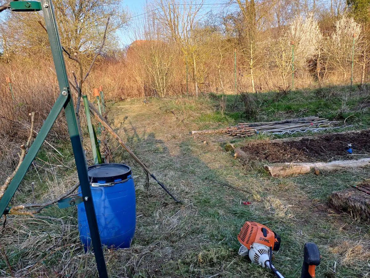 Vue réelle en Moselle d'une débroussailleuse au premier plan, devant un potager en préparation. Sur le côté droit, des planches de culture déjà surélevées avec des rondins de bois et du terreau noir sont prêtes pour la plantation. Cette photo illustre concrètement comment installer un petit potager débutant sans bêcher, en gérant la végétation existante avant de créer une zone fertile.